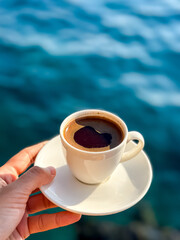 Female hand holding white cup with Turkish coffee or espresso. Sea water on a background. Coffee break. Time for coffee. Holidays.