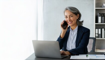 Happy smiling mature middle aged business woman, 40s professional lady executive manager talking on the phone making business call on cellphone at work in office using laptop compute