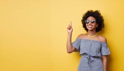 Young happy African gen z girl in sunglasses standing at yellow wall pointing up at copy space. Smiling Black woman presenting promo on summer day advertising shopping sale offers, vertical photo