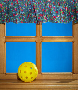 Yellow Pickleball in window setting with curtain and blue background.