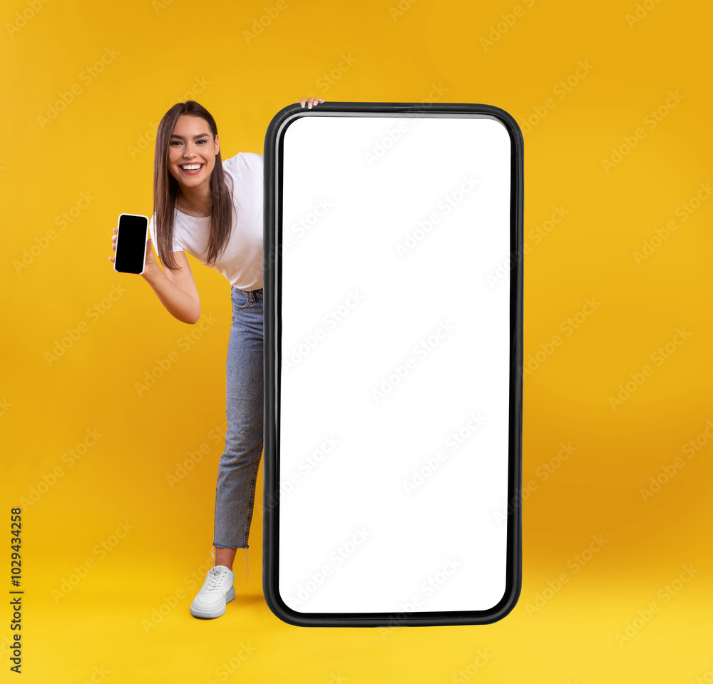 Poster full body length of cheerful woman peeking out standing behind big smartphone with white blank scree