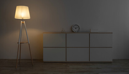 White furniture, clock, glowing lamp on floor in evening on gray wall background in office or living room, nobody, copy space. Modern scandinavian home interior, minimalist design, furniture in dark