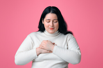 Fototapeta premium Gratitude concept. Thankful overweight woman keeping both palms on chest, expressing appreciation on pink studio background with copy space
