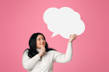 Pensive plus size woman holding and looking at blank speech bubble over her head, standing on pink...