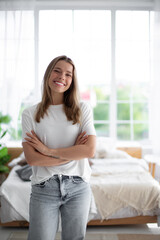 Cute woman smiles confidently with arms crossed, wearing white t-shirt and jeans in a bright, airy bedroom. The room features large windows, natural light, greenery, and soft bedding, evoking comfort