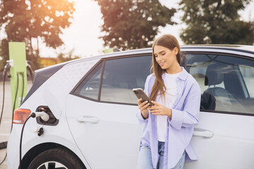 Young Woman Smiling While Using Smartphone By Electric Car At Charging Station Outdoors