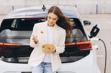 Naklejka premium Young Woman Enjoying Healthy Lunch While Charging Electric Car in Urban Parking Lot