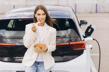 Naklejka premium Young Woman Enjoying Healthy Lunch While Charging Electric Car in Urban Parking Lot