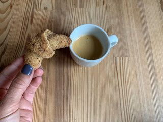 Minimalist wooden table with white cup of fresh espresso and female hand holding small croissant, perfect for cozy breakfast or cafe related images