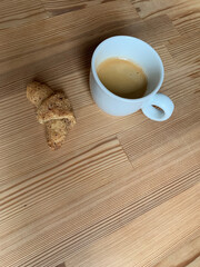 Minimalist wooden table with white cup of fresh espresso and small croissant next to it, perfect for cozy breakfast or cafe related images