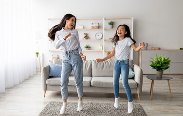 Playful korean mother and pretty daughter dancing to music and jumping at home. Asian mom and her kid enjoying favorite song together, fooling around, having fun in living room interior