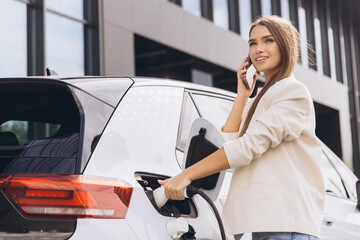 Woman Charging Electric Car While Talking on Phone Outdoors