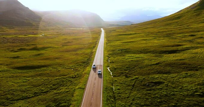Aerial shot of white electric car traveling through breathtaking sunny landscape in the Highlands Scotland