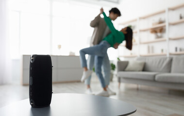 Millennial Asian couple dancing to popular music at home, selective focus on portable wireless speaker on table, copy space. Young family moving to their favorite song, using modern device