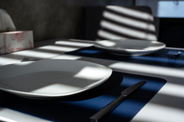 A white table with dark blue place mats, two plates, cutlery, and a box of tissues. Sunlight from...