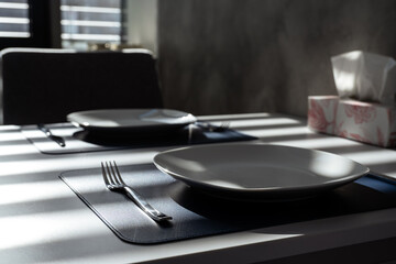 A dining table with a navy blue place mat, white plate, and silver cutlery. Sunlight through window blinds creates shadow patterns on the scene.