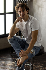 Young sexy handsome fit stylish man in a white shirt looking at the camera sitting on a chair in the studio. Portrait in a studio, casual clothes, modern style