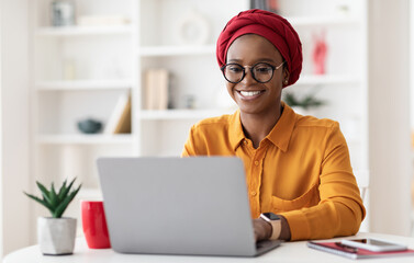 Cheerful millennial black woman entrepreneur working from home, wearing red headscarf and...