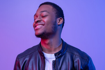 Closeup Shot Of Satisfied Black Guy In Leather Jacket Standing With Closed Eyes Over Purple Studio Background, Pleased Handsome African American Man Posing Under Neon Lighting, Copy Space