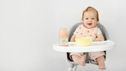 Adorable baby girl sitting in high chair and smiling at camera, eating from plate, panorama with free space. Baby food concept