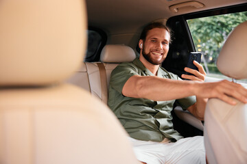 Smiling man sitting in the backseat of a car, holding a smartphone and looking at the screen