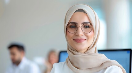 Confident young professional woman wearing glasses and a hijab, smiling in a modern office environment with colleagues in the background.