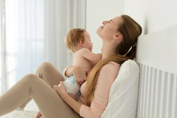 Tired crying young mother suffering from experiencing postnatal depression, sitting on bed and holding baby on hands