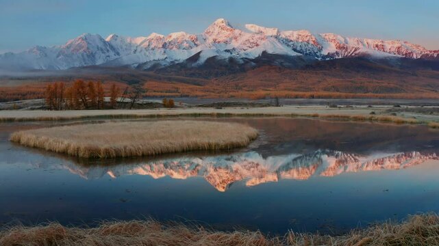 Experience the serene beauty of Mountain Altai captured by drone in late autumn. Snow capped peaks and golden trees reflect perfectly in calm water, showcasing nature stunning colors and tranquility