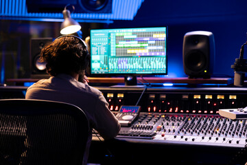 Audio engineer mixes tracks to create a new song for his album, editing tunes with digital software interface and mixing console in control room. Sound technician works on post production.