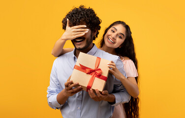 Pretty indian woman covering her boyfriend's eyes, holding wrapped gift box and greeting him with...