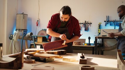 Carpenter in assembly shop starting restoration project on lumber block, applying lacquer on surface. Woodworking specialist coloring wood plank with paintbrush, doing handwork in studio, camera B
