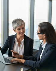 Fototapeta premium Two busy professional business people working in office with laptop computer. Mid aged mature Latin female executive manager talking to male colleague having conversation sitting at workplace