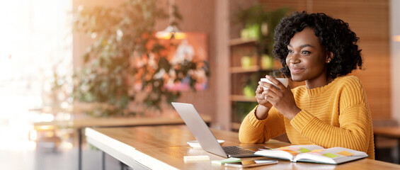 Dreamy african girl dinking tea at cafe while looking for job online, using laptop and taking...