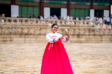 Naklejka premium Happy young Latina wearing a pink and white hanbok, standing in front of Gyeongbokgung Palace, taking selfie in Seoul, South Korea.