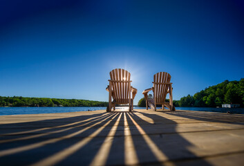 Naklejka premium At sunrise, a pair of empty Adirondack chairs sit peacefully on a dock overlooking a lake in Muskoka, Ontario, Canada. The soft morning light creates long shadows on the weathered wooden pier. 