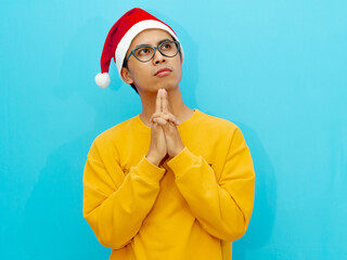 Serious Asian man wearing a Christmas hat, looking thoughtful and gazing upward, isolated against a blue background.