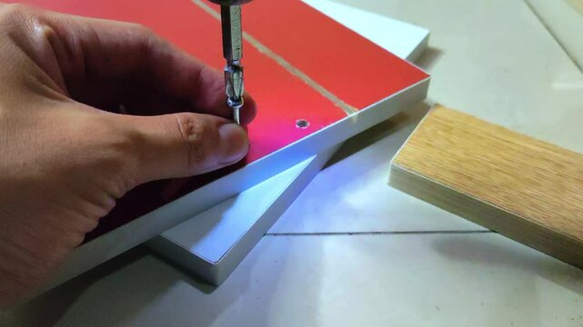 a man's hand installing a minifix screw into a red wooden board using a electric screwdriver drill machine.