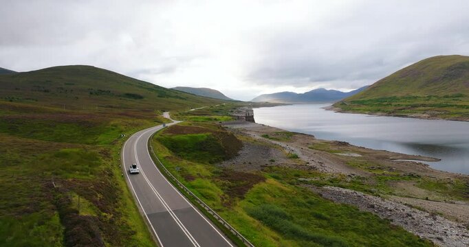 Aerial view of white electro car driving through epic landscape of Scottish hills and fields near cinematic calm lake