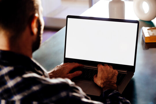 Over shoulder view of a young business man using computer laptop in front of an blank white computer screen in his home living room. Photo ready for mockup.