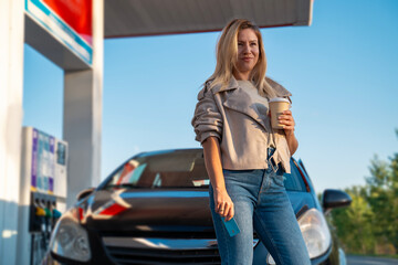 Confident attractive woman on gas station standing in front of the car with cup of coffee in her...