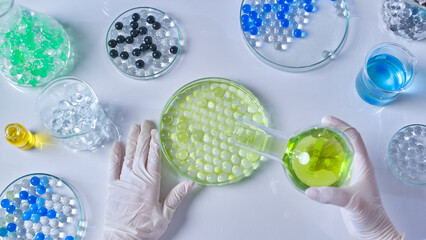 Top shot photography at white laboratory table top, a scientist is pouring yellow essence from a boiling flask into glass petri dish of gel ball placed on table, which surrounded by other lab utensils