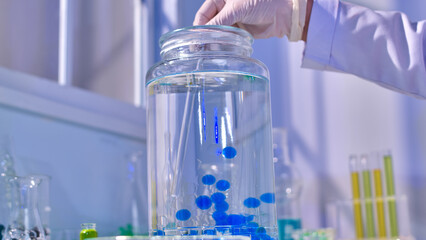 Creative template photo for advertising pic design, a scientist's hand holds glass rod to stir hydrogel balls and water in large glass vase. Photo was taken from below view, designing space