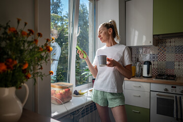 Middle-aged woman drinking coffee, holding mug, using phone and scrolling social media, distraction on real life, standing at kitchen. Relaxed awaked female using smartphone spending lazy start day. 