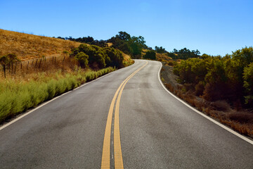 Mt. Hamilton Road winds through a dry, rural landscape with grassy hills and bushes under a clear blue sky