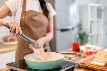 A woman holding tongs, flipping a slice of bacon in a pan while making her breakfast in the kitchen.