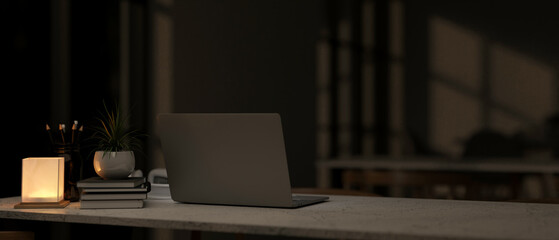 A back view of a laptop on a table in a modern dark room at nighttime, illuminated by a table lamp.
