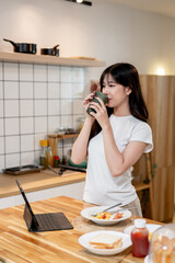 A beautiful young Asian woman is drinking water after having breakfast in the kitchen.