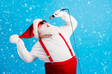 Photo of old man grey beard hold mic open mouth sing song wear santa claus x-mas costume headwear sunglass suspenders white shirt gloves isolated blue color background