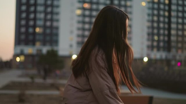 A girl in a peach jacket and black trousers is seen skating towards a bench in an urban park. After sitting down, she rests her hand on the bench, with her face turned slightly away from the camera