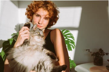 portrait red-haired curly young woman with pet gray fluffy cat in arms, lit by bright sun